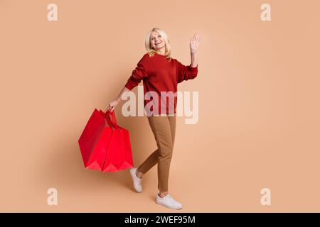 Foto in voller Länge von traumhafter aufgeregter Dame gekleidet roter Pullover, der Käufer hält, die hoch aussehenden leeren Raum winken, isolierter beigefarbener Hintergrund Stockfoto