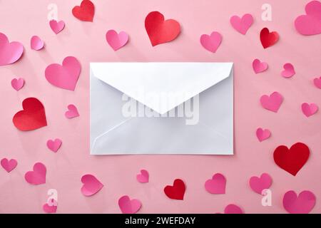 Weißer Umschlag in der Mitte auf rosafarbenem Hintergrund mit Papierausschnitten aus rosa und roten Herzen. Draufsicht. Stockfoto