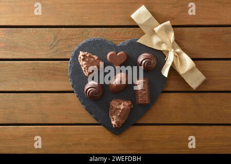 Schokoladensortiment auf herzförmigem Schieferteller mit goldener Schleife in der Mitte auf Holzlattentisch. Draufsicht. Stockfoto