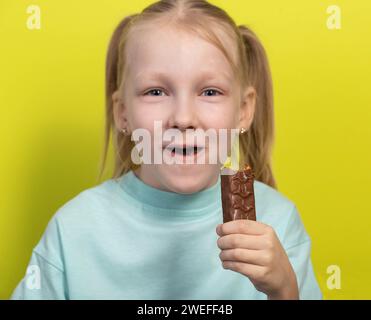 Ein schönes, fröhliches Mädchen mit weißen Haaren von sieben Jahren isst eine Schokoladentafel auf gelbem Hintergrund. Kinder lieben süßes Junk Food, Horizo Stockfoto