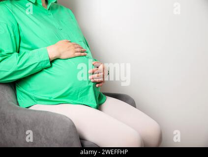 Ein schwangeres Mädchen in einer grünen Bluse sitzt auf einem Stuhl und zählt regelmäßige Wehen vor Beginn der Geburt und einem Ausflug in die Entbindungsklinik. Kopieren Stockfoto