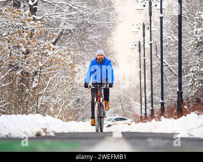Ein bärtiger Mann fährt im Winter auf einem Radweg in der Stadt. Aktiver Lebensstil. Ein Typ in einer blauen Jacke auf einem Fahrrad Stockfoto