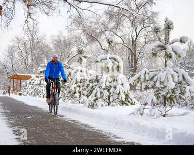 Ein fröhlicher Mann fährt mit dem Fahrrad in einem Winterpark zwischen schneebedeckten Bäumen. Bärtiger Radfahrer in blauer Jacke. Aktiver Lebensstil Stockfoto