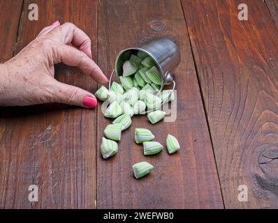 Frauenhand nimmt Minzbonbons aus silbernem Mini-Eimer, die grün und weiß gestreifte Bonbons auf braunen Holztisch verteilen Stockfoto