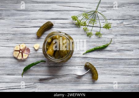 Gurken in Dosen in einem offenen Glas, eine eingelegte Gurke wird auf eine Gabel gestochen und liegt in der Nähe auf einem grauen hölzernen Hintergrund, Blick von oben. Stockfoto