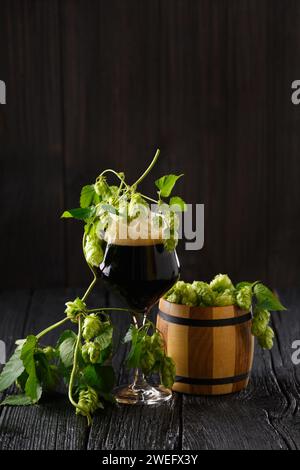 Glasbecher mit dunklem Bier, Hopfenzweigen und einem Holzfass mit Hopfen auf dunklem Holzhintergrund. Brautraditionen. Oktoberfest. Bierfest. Stockfoto