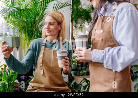 Besitzer mittelalter blonde Frau, die sich um die Pflanzen im Topf kümmert. Ihre Assistentin hilft beim Inventar in einem gemütlichen Pflanzenladen. Stockfoto