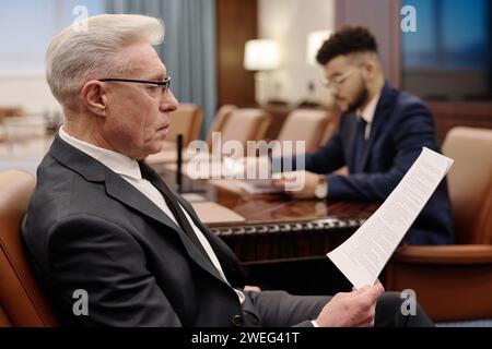 Reifer Geschäftsmann liest Dokumente im Sitzungssaal, sein junger Kollege arbeitet an Hintergründen Stockfoto