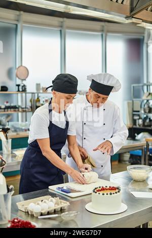 Attraktiver, reifer Koch in Schürze und Haube, der neben ihrem Chefkoch, dem Süßwaren, mit Teig arbeitet Stockfoto