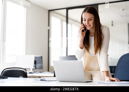 Eine junge Geschäftsfrau in dynamischer Arbeitspose, die am Telefon spricht und ihren Laptop benutzt, was ihre Fähigkeit unterstreicht, mehrere Aufgaben in einer schnelllebigen Arbeitsumgebung effizient zu bewältigen Stockfoto