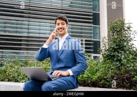 Ein junger hispanischer Geschäftsmann führt ein Gespräch, während er an seinem Laptop arbeitet. Er sitzt draußen im Grünen in der Nähe eines Bürokomplexes und blickt freundlich in die Kamera Stockfoto