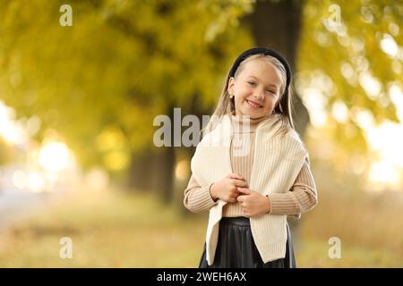 Kindertag. Fröhliches kleines Mädchen 5-6 Jahre alt posiert und lächelt draußen vor der Kamera und steht am Baum im Herbstpark. Stilvoller Zartpressdruck Stockfoto