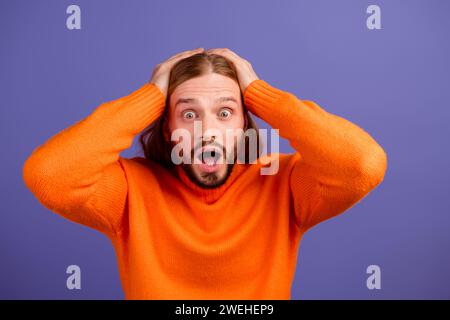 Foto eines jungen Mannes, der einen hellorangefarbenen Pullover trägt, überrascht den berührenden Kopf. Unglaubliche wtf-Reaktion isoliert auf violettem Hintergrund Stockfoto