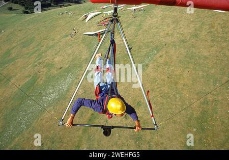 Aus Sicht des Piloten hängt ein junger Mann in einem Drachenflieger und fliegt über den Startplatz, wo andere Drachenflieger zu sehen sind, Monte Cucco, Umbrien, Ita Stockfoto