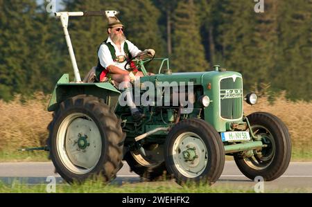 Ein Mann in Lederhosen, mit Vollbart, Hut, Sonnenbrille und traditioneller Tracht sitzt auf einem alten grünen MANN Traktor und fährt durch die Landschaft ne Stockfoto