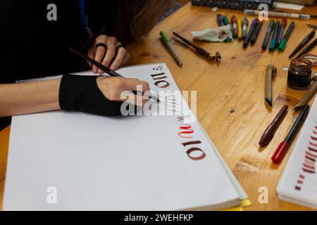 Eine erfahrene Kalligrafin übt das Schreiben und Zeichnen von Schriften im Stil der Renaissance in einem Skizzenbuch. Auf dem Holztisch gibt es Markierungen und Farben Stockfoto