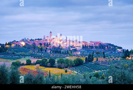 Blick auf die Stadt San Gimignano auf einem Hügel am frühen Morgen zur blauen Stunde mit bewölktem Himmel im Herbst, überall sind Zypressen, Olivenhaine und Weinberge, Stockfoto