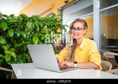 Lächelnde junge Freiberuflerin mit Laptop am Kaffeetisch. Frau in gelber Jacke mit blonden Haaren, die neben grüner Pflanze auf der Terrasse des Cafés sitzt. Konzept für Fernarbeit. Stockfoto