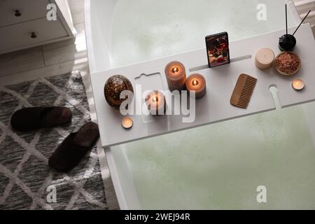 Weißes Holztablett mit Smartphone, brennenden Kerzen und Spa-Produkten auf der Badewanne im Bad, Blick über dem Zimmer Stockfoto