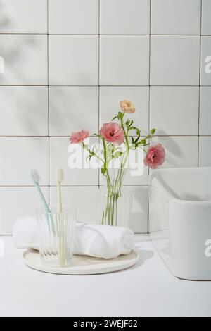 Schönheitsfoto mit Badezimmerkonzept. Zahnbürsten im Glasbecher, weißes Handtuch auf runden Geschirr, eine Ecke des Waschtisches und rosa Blumenvase auf Fliesen W Stockfoto