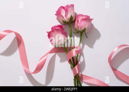 Zarte Rosen, die um ein zartes rosafarbenes Band auf weißem Hintergrund gewickelt sind. Flache Komposition mit schöner Blume für Werbung für Kosmetik oder Design Stockfoto