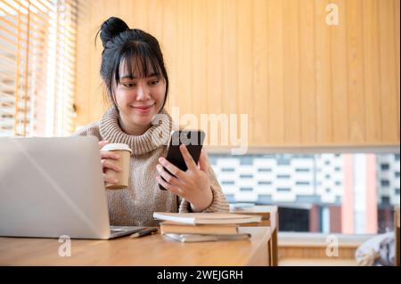 Eine positive asiatische Frau in einem gemütlichen Pullover schlürft Kaffee und liest SMS auf ihrem Handy, während sie in einem Café arbeitet. People und lifestyl Stockfoto
