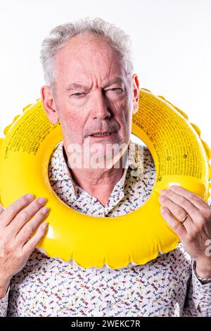 Dieses Bild zeigt einen älteren Mann mit einem skeptischen oder unsicheren Ausdruck, während er einen gelben Schwimmring um den Hals hält. Der Kontrast zwischen dem hellen, verspielten Objekt und seinem dubiosen Ausdruck erzeugt einen humorvollen und ansprechenden Moment, der vielleicht das Zögern widerspiegelt, das manche bei neuen Aktivitäten oder der komödiantischen Seite des Sommerurlaubs empfinden. Das Bild kann die hellere Seite des Alterns symbolisieren und ermutigen, trotz anfänglicher Vorbehalte einen Sinn für Humor und die Bereitschaft, Spaß zu machen. Senior Skeptical Look mit Swim Ring. Hochwertige Fotos Stockfoto