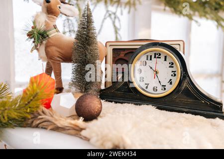 Helle Komposition des Innenraums des Weihnachtszimmers mit stilvollem Kranz, Uhr, kleinem Weihnachtsbaum, Ball auf dem Hintergrund des gefrorenen Fensters am sonnigen Wintertag. Stockfoto