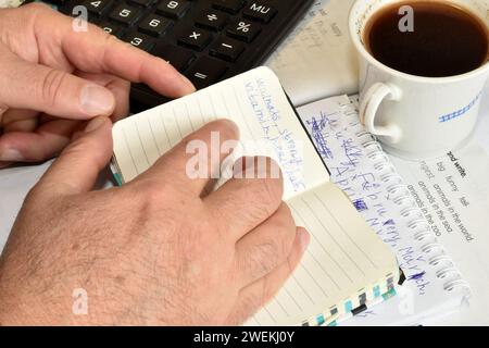 Das Bild zeigt einen, der Hände eines Mannes durch ein Notizbuch blättert. Ein Mann sitzt an einem Tisch, auf dem sich eine Tasse Kaffee, ein Taschenrechner und Papiere befinden. Stockfoto