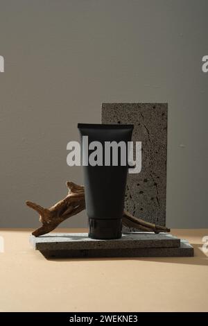 Eine schwarze Plastikflasche ohne Etikett und kleinen getrockneten Zweig auf grauen Steinen auf dunklem Hintergrund. Minimalistischer Kunsthintergrund für Mockup, Kosmetikprofi Stockfoto