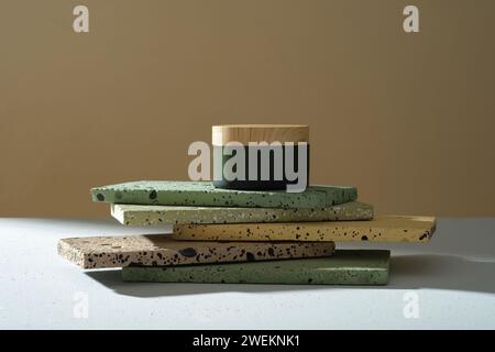 Grüne Gläser ohne Etikett mit Holzdeckel auf bunten rechteckigen Ziegeln, die auf braunem Hintergrund gestapelt sind. Modell für Kosmetik, Feuchtigkeitspflege, Maske oder Stockfoto