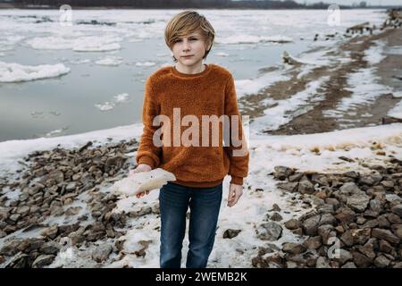 Junge vor Teenager am eisigen Ufer, der Eisbrocken hält Stockfoto