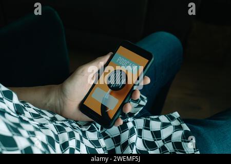 Nahaufnahme eines jungen Mannes in Freizeitkleidung, der auf einem bequemen Stuhl sitzt und einen falschen Podcast in seinem Smartphone hört, der den Text-Podcast liest Stockfoto