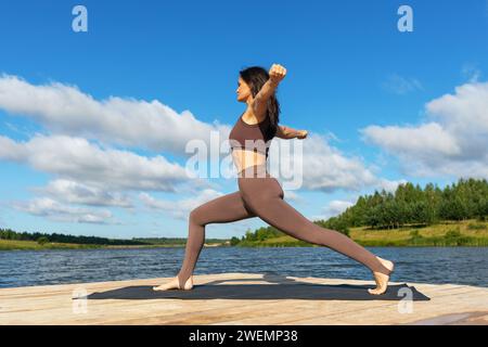 Attraktive Frau, die eine Variation von Virabhadrasana-Übungen, Kriegerstellung, Training in Leggings und einem kurzen Top macht, während sie am Ufer steht Stockfoto