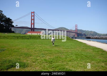 Die Golden Gate Bridge ist eine Hängebrücke, die das Golden Gate überspannt, mit einer jungen Frau, die auf Gras unter ihr läuft und die Brücke in der Ferne liegt. Stockfoto