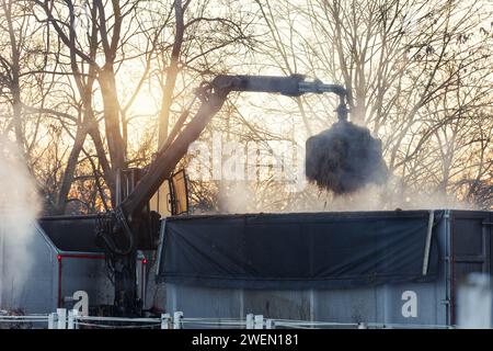 Moderner LKW Kranarm Ladeanhänger Haufen Dung Strohdünger von Pferd Kuh Bauernhof Hof Landschaft warmer Sonnenuntergang Hintergrund. Dampfrauchen Stockfoto