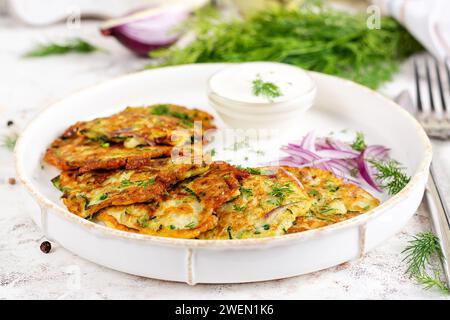 Zucchini-Krapfen mit roten Zwiebeln, Knoblauch und Kräutern. Vegetarische Zucchini-Pfannkuchen und Sauerrahm auf weißem Tisch. Stockfoto