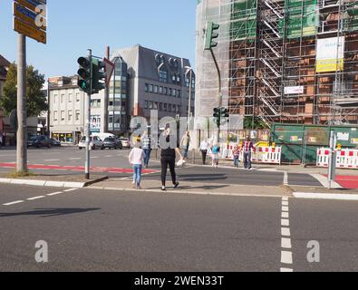 Fußgänger und Kinder überqueren die Hauptstraße an der Kreuzung während der Grünphase an der Ampel in der Stadt an einem sonnigen Tag mit Gerüstkirche Stockfoto