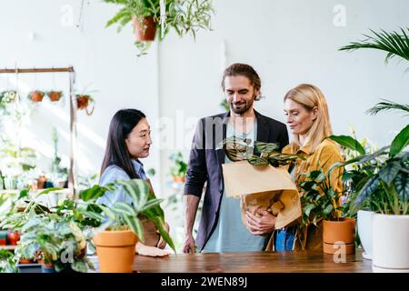 Blumenladen. Asiatische Floristin, die mit männlichen und weiblichen Kunden spricht. Fröhliche Pärchen, die Pflanzen kaufen. Frau, die an der Theke steht und Stockfoto