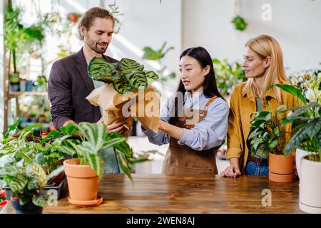 Erwachsene asiatische Arbeiterin Gärtnerin mit Schürze, Verpackung in Papier und Verkauf Haus Pflanztöpfe für glückliche europäische Paare im Blumenladen. Florist Stockfoto