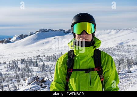 Nahaufnahme eines Skifahrers oder Snowboarders in Sportausrüstung, schneebedeckte Berge im Skigebiet. Leuchtendes, säuregrünes Outfit: Warme Anzugsjacke, Stockfoto