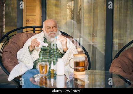 Der alte Mann mit Kälte ist in eine flauschige Decke auf der Veranda gewickelt Stockfoto