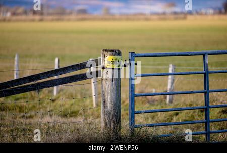 Holzpfosten mit einem elektrischen Zaunschild in landwirtschaftlicher Umgebung, mit einem verzinkten Tor auf der einen Seite und einem Zaun auf der anderen Seite Stockfoto