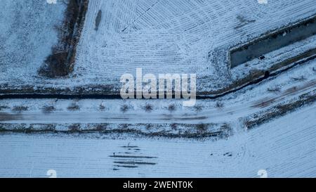 Dieses Luftbild zeigt einen fesselnden Blick auf eine frostige landwirtschaftliche Landschaft. Das Bild zeigt das grafische Zusammenspiel zwischen den Kurven einer schneebedeckten Straße und den geometrischen Mustern der umliegenden Felder, wodurch ein natürlicher Flickenteppich aus kontrastierenden Texturen entsteht. Die Spuren des Schnees heben die landwirtschaftlichen Linien und die Reste der Ernte hervor, während ein Hauch von Grün hindurchblickt und auf das Leben darunter hinweist. Frosty Air Patchwork. Hochwertige Fotos Stockfoto