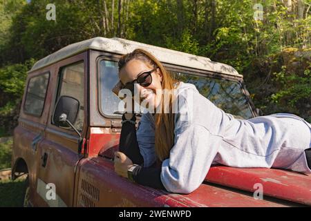 Lächelnde Frau, die auf der Motorhaube eines alten roten Geländewagens liegt, der mitten im Wald der Pyrenäen geparkt ist Stockfoto