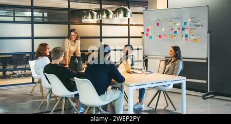 Geschäftsleute in einem Sitzungssaal führen ein kreatives Gespräch und tauschen sich mit einer selbstbewussten Führungskraft aus, die das Meeting unterstützt. Das glückliche Team repräsentiert Stockfoto