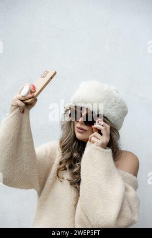 Eine stilvolle Frau in einem cremigen Off-Shoulder-Pullover und einer luxuriösen Pelzmütze macht ein Selfie, das Eleganz und Wintercharme ausstrahlt. Stockfoto