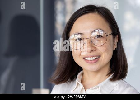Entzückende asiatische Frau mit kurzen dunklen Haaren und Brille, die in die Kamera blickt und lächelt. Junge, fröhliche Arbeiterin in weißem Hemd, die im modernen Büro steht und positive Emotionen ausdrückt. Stockfoto