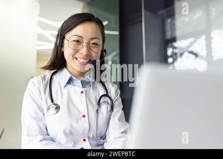 Freundliche asiatische Sanitäterin in Brille, die Videoanrufe auf Laptop während sie im Büro sitzt. Ärztin mit Uniform und Headset, die während des Online-Meetings auf den Computerbildschirm schaut und lächelt. Stockfoto