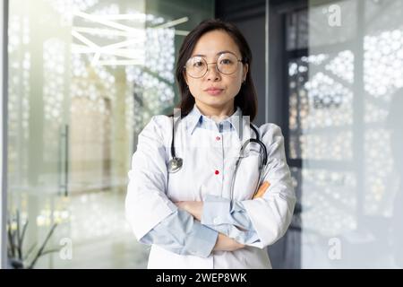 Konzentrierte asiatische Ärztin mit weißem medizinischem Gewand und Stethoskop, stehend mit überkreuzten Armen und Blick in die Kamera. Selbstbewusste Therapeutin bereit, professionelle Hilfe im Krankenhaus zu leisten. Stockfoto